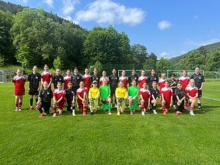 Gruppe junger Fußballspielender in roten und schwarzen Trikots auf Rasen, in Reihen aufgestellt, mit Toren, Bäumen und blauem Himmel im Hintergrund.