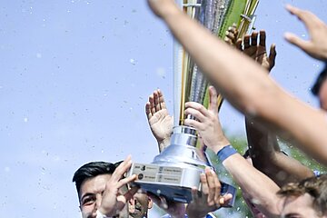Das Bild zeigt eine Gruppe von Menschen, die gemeinsam einen großen silbernen Pokal hochhalten. Der Pokal hat eine breite Basis und verjüngt sich nach oben hin, mit mehreren Griffen oder Vorsprüngen in der Nähe des oberen Teils. Der Hintergrund ist unscharf und zeigt eine Außenumgebung mit klarem blauen Himmel und etwas Grün. Foto: Sandra Ritschel