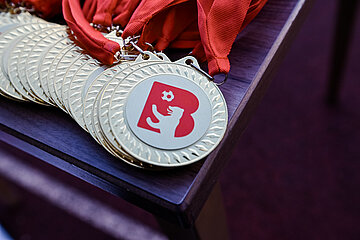 Das Foto zeigt eine Nahaufnahme mehrerer goldener Medaillen, die auf einem dunklen Holztisch liegen. Jede Medaille ist an einem roten Band befestigt. Auf der Vorderseite der Medaillen ist ein rotes Logo zu sehen: Es zeigt einen stilisierten Buchstaben „B“ sowie eine Figur, die gerade einen Fußball tritt. Die Medaillen sind ordentlich nebeneinander platziert und glänzen im Licht.