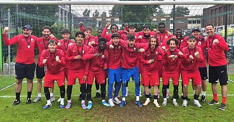 Youth soccer team in red jerseys posing on grass in front of goal and fence, several wearing medals and raising their fists.