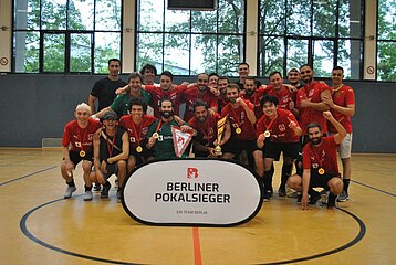 Eine Fußballmannschaft in roten Trikots feiert in einer Sporthalle den Berliner Pokalsieg. Vor ihnen steht ein Schild mit „Berliner Pokalsieger“. Foto: BFV