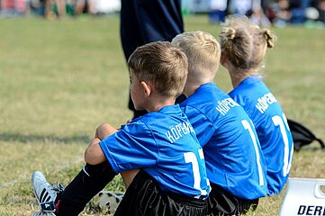 Das Foto zeigt drei Kinder, die auf dem Boden sitzen und offenbar eine Pause während eines Fußballspiels machen. Sie tragen blaue Trikots mit weißen Rückennummern und dem Schriftzug „Köpenick“ auf dem Rücken. Alle drei haben schwarze Shorts und Fußballschuhe an. Die Kinder sitzen nebeneinander auf einer Wiese, die als Spielfeld dient, und schauen in dieselbe Richtung, vermutlich zum laufenden Spiel. Im Vordergrund ist ein Teil eines Schildes mit der Aufschrift „DER“ zu sehen. Der Hintergrund ist unscharf, aber es sind weitere Personen und Spielfeldbegrenzungen erkennbar, was auf eine sportliche Veranstaltung im Freien hinweist.