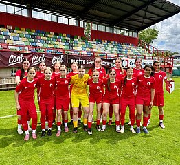 Gruppe junger Fußballspielerinnen in roten Trikots steht auf Rasen vor Tribüne mit bunten Sitzen, mittig Person im gelben Torwartoutfit