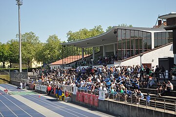 Ein großes, offenes Stadion, nämlich das Stadion Lichterfelde, ist gut besucht. Die Zuschauertribünen sind überdacht und mit Glasfenstern versehen. Viele Menschen sitzen oder stehen dicht gedrängt und blicken auf das Spielfeld. Man sieht eine blaue Laufbahn. Entlang der Brüstung vor den Tribünen hängen mehrere Banner. Im Hintergrund sind grüne Bäume zu sehen, was darauf hinweist, dass sich das Stadion im Freien befindet.