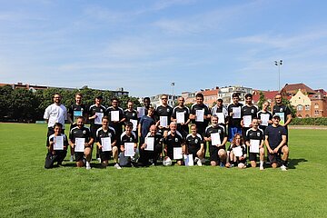 Eine Gruppe von Personen steht auf einem grünen Sportfeld. Alle tragen schwarze Sportkleidung und halten Zertifikate in den Händen. Im Hintergrund sind Bäume und Gebäude zu sehen, der Himmel ist klar und blau.