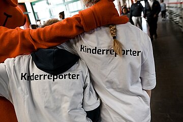 Auf dem Foto stehen zwei Kinder nebeneinander, beide tragen weiße T‑Shirts mit der Aufschrift „Kinderreporter“ auf dem Rücken. Eine große Figur mit orangefarbenem Kostüm legt einem der Kinder einen Arm freundschaftlich über die Schulter. Die Szene spielt in einem hellen Innenraum, in dem weitere Personen im Hintergrund zu sehen sind. Die Atmosphäre wirkt lebhaft und fröhlich.