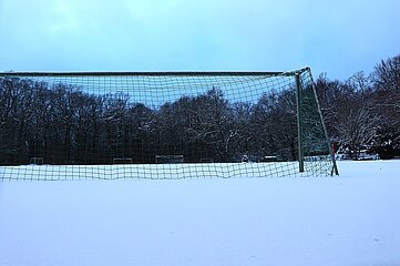 Das Foto zeigt ein einzelnes Fußballtor im Vordergrund, aufgenommen aus einer niedrigen Perspektive knapp über dem Boden. Der gesamte Boden ist von einer dicken, unberührten Schneeschicht bedeckt, die eine gleichmäßig weiße Fläche bildet. Das Fußballtor besteht aus einem Metallrahmen mit einem grünen Netz, das leicht durchhängt. Der Schnee liegt auch am unteren Rand des Netzes und berührt teilweise die Pfosten. Im Hintergrund erstreckt sich eine dichte Baumreihe, deren Äste ebenfalls mit einer dünnen Schicht Schnee bedeckt sind. Die Bäume wirken winterlich kahl. Der Himmel darüber ist hellblau bis grau und wirkt wolkig, was der Szene eine ruhige und kalte Atmosphäre verleiht. Das Foto vermittelt insgesamt den Eindruck eines komplett verschneiten, stillen Sportplatzes im Winter, ohne sichtbare Menschen oder Tiere.