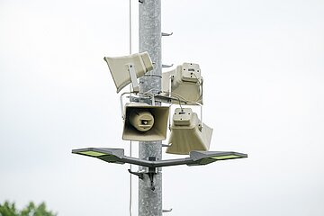 Auf dem Bild ist ein metallischer, senkrechter Mast im Außenbereich zu sehen, an dem mehrere beigefarbene Lautsprecher befestigt sind. Die Lautsprecher sind trichterförmig und in unterschiedliche Richtungen ausgerichtet, sodass sie Schall in verschiedene Richtungen abgeben können; Kabel verbinden die einzelnen Geräte miteinander. Unterhalb der Lautsprecher ist eine Straßenlaterne mit zwei flachen, rechteckigen Leuchten angebracht, die nach links und rechts zeigen. Der Hintergrund ist hell und weitgehend einheitlich, vermutlich ein bewölkter Himmel, am unteren Bildrand sind unscharf grüne Baumkronen erkennbar.