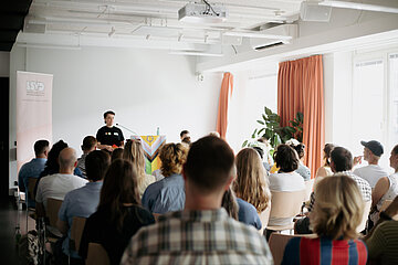 Person at the lectern in front of a seated audience in a bright room; banner, plant, curtains and ceiling technology visible. Photo: LSVD Queer Diversity Association Berlin-Brandenburg 