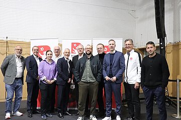 Das Bild zeigt eine Gruppe von neun Personen, die in einer Halle zusammenstehen. Sie bilden eine lockere Reihe und stehen vor zwei großen, weißen Roll-Up-Bannern mit dem roten Logo des Berliner Fußball-Verbands (BFV). Das Logo besteht aus einem stilisierten „B“ mit einem Fußballmotiv. Die Personen tragen unterschiedliche Kleidung: Einige sind in formellen Anzügen mit Hemd und Krawatte gekleidet, andere in Freizeitkleidung wie Jeans, Hemd oder Pullover. Eine Person trägt eine weiße Sportjacke mit dem BFV-Logo auf der Brust. Die Schuhe variieren von eleganten Lederschuhen bis zu sportlichen Sneakern. Der Hintergrund ist schlicht: eine helle Wand mit einem Netz an der oberen Hälfte, vermutlich Teil einer Sporthalle. Rechts im Bild ist ein schwarzer Lautsprecher auf einem Stativ zu sehen. Der Boden ist dunkel, wahrscheinlich Teppich oder Gummibelag. Die Beleuchtung ist hell und gleichmäßig, was auf eine Innenaufnahme mit künstlichem Licht hinweist. Die Szene vermittelt den Eindruck eines offiziellen Gruppenfotos, vermutlich nach einer Wahl oder einem formellen Anlass.
