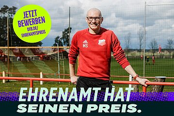 Auf dem Bild steht ein lächelnder Mann auf einem Fußballplatz. Er trägt eine rote Trainingsjacke mit dem Aufdruck „MS“ sowie ein Vereinslogo. Im Hintergrund sind ein Fußballfeld, Tore und einige spielende Personen zu sehen. Der Mann lehnt mit einer Hand entspannt an einem rot‑orangenen Geländer. Links oben befindet sich ein großer gelber Kreis mit der Aufschrift: „Jetzt bewerben – dfb.de/ehrenamtspreis“. Unten im Bild steht in großen, hellen Buchstaben: „Ehrenamt hat seinen Preis.“ Der Gesamtstil wirkt freundlich und motivierend.