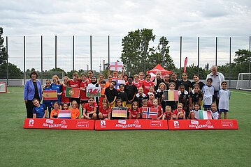 Auf dem Bild ist eine große Gruppe von Menschen auf einem Fußballfeld zu sehen – darunter Kinder und Erwachsene. Sie stehen eng beieinander und posieren gemeinsam für ein Gruppenfoto. Viele von ihnen halten bunte Nationalflaggen in den Händen, darunter die Flaggen von Spanien, Portugal, Wales, Deutschland, Norwegen und Italien. Im Hintergrund sind rote Absperrungen mit der Aufschrift „Alle kicken mit!“ und „Berlin“ zu erkennen. Hinter den Absperrungen stehen Bäume und ein Zaun, die das Spielfeld begrenzen. Die Stimmung wirkt gemeinschaftlich und festlich.