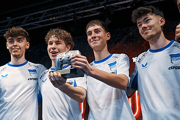 Auf dem Foto stehen vier Personen eng beieinander auf einer Bühne oder Eventfläche. Sie tragen weiße Trikots mit blauen Details und einem Vereins‑ oder Teamlogo auf der Brust. Alle stehen Schulter an Schulter, als würden sie gemeinsam einen Erfolg feiern. Im Mittelpunkt des Bildes halten zwei Personen gemeinsam eine Trophäe oder einen Preis, der wie ein Gaming‑ oder E‑Sport‑Award aussieht. Ihre Hände umfassen den Sockel des Pokals, den sie stolz nach vorne präsentieren. Der Hintergrund zeigt eine große, farbintensive Bühnenwand mit LED‑Grafiken, die an ein professionelles E‑Sport‑Event erinnert. Über ihnen ist Bühnentechnik zu erkennen, wie Scheinwerfer und Metallträger. Die Szene vermittelt Teamgeist, gemeinsames Feiern und den Moment eines sportlichen oder spielerischen Erfolgs.