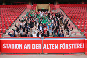 Das Foto zeigt eine große Gruppe von Menschen auf den Tribünen eines Fußballstadions. Die Personen sitzen und stehen auf roten Sitzreihen, die sich stufenförmig nach oben erstrecken. Viele der Anwesenden heben beide Arme nach oben und zeigen offene Hände. Die Personen tragen unterschiedliche Kleidungsstücke, unter anderem Jacken, Pullover, Hemden und Schals in verschiedenen Farben, darunter Grün, Schwarz, Grau und Blau. Einige Personen tragen Brillen, andere Kopfbedeckungen. Die Gruppe wirkt altersmäßig vielfältig. Im unteren Vordergrund befindet sich eine rote Brüstung mit weißer, gut lesbarer Schrift. Darauf steht: „STADION AN DER ALTEN FÖRSTEREI“. Die Schrift ist groß und kontrastreich gestaltet. Im Hintergrund sind weitere Reihen roter Sitzplätze zu sehen, die überwiegend leer sind, sowie Treppenaufgänge mit hellen Stufen und roten Geländern. Die Architektur des Stadions ist klar strukturiert und symmetrisch. Die Szene ist bei Tageslicht aufgenommen, die Beleuchtung ist gleichmäßig, ohne starke Schatten. Das Bild vermittelt den Eindruck einer gemeinsamen Veranstaltung oder eines Gruppentreffens an einem bekannten öffentlichen Ort. Die Menschen sind gleichberechtigt nebeneinander platziert und gemeinsam Teil der Szene.