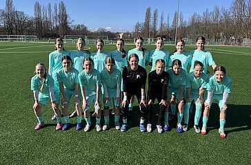 Eine Fußballmannschaft steht auf einem grünen Kunstrasenplatz, umgeben von kahlen Bäumen unter einem klaren blauen Himmel. Die Gruppe trägt einheitliche türkisfarbene Trikots, kurze Hosen und Stutzen; zwei Personen in der vorderen Reihe tragen dunklere Trainingskleidung. Die vordere Reihe kniet oder hockt, während die hintere Reihe steht. Im Hintergrund sind weitere Fußballfelder, weiße Linienmarkierungen und ein hoher Flutlichtmast zu sehen. Das Team wirkt ordentlich aufgestellt, als würde ein offizielles Mannschaftsfoto aufgenommen.