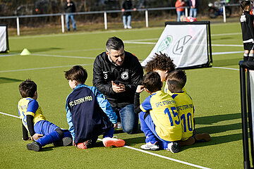 Auf dem Foto ist eine Gruppe von fünf Kindern und einer erwachsenen Person auf einem Fußballplatz zu sehen. Die Kinder tragen bunte Fußballtrikots – einige in Gelb-Blau, andere in Blau-Schwarz – mit der Aufschrift „SFC STERN 1900“ auf dem Rücken. Sie sitzen oder knien auf grünem Kunstrasen und hören aufmerksam der erwachsenen Person zu, die sportlich gekleidet ist und ihnen offenbar etwas erklärt oder zeigt. Die Szene wirkt wie eine Trainings- oder Spielbesprechung. Im Hintergrund sind weitere Menschen sowie ein kleines Fußballtor mit sichtbaren Logos zu erkennen. Die Atmosphäre ist lebendig und gemeinschaftlich.