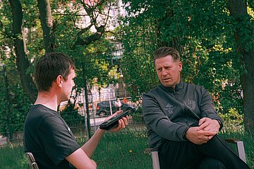 Zwei Personen sitzen draußen im Gespräch. Die linke Person hält ein Smartphone oder Aufnahmegerät in der Hand und richtet es auf ihr Gegenüber, das entspannt mit verschränkten Händen zuhört.