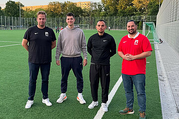 Das Foto zeigt vier Personen, die nebeneinander auf einem Fußballfeld stehen. Sie blicken direkt in die Kamera und wirken präsent und aufmerksam. Die Personen unterscheiden sich in Kleidung, Körperbau und Erscheinung, was auf eine vielfältige Gruppe hinweist.  Die erste Person von links trägt ein schwarzes T-Shirt mit einem weißen Logo und eine dunkle Hose. Die zweite Person hat einen grauen Kapuzenpullover und eine dunkle Hose an. Die dritte Person ist ganz in Schwarz gekleidet – mit einem schwarzen Oberteil und einer schwarzen Hose. Die vierte Person trägt ein rotes T-Shirt mit einem weißen Logo und eine blaue Jeanshose. Im Hintergrund sind weitere Menschen zu sehen, die sich auf dem Fußballfeld bewegen oder spielen. Das Feld ist von einem hohen Zaun umgeben, und dahinter stehen einige Bäume. Die Szene spielt sich im Freien bei Tageslicht ab.