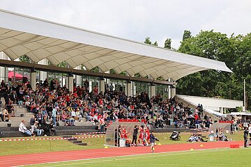 A large, covered stadium stand is almost completely filled with spectators. The stand stretches from left to right and consists of several rows of concrete seats. In the foreground is a red running track on which individual children in sportswear and supervisors stand or walk. At the edge of the running track is a transparent players' bench. Glass fronts and white supporting pillars can be seen behind the stands, with a bright, curved roof stretching above them. Green trees frame the stadium on the right and in the background. The photo shows a well-attended sporting event in daylight.