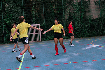 Mehrere Personen spielen auf einem Outdoor-Sportplatz; zwei Personen in gelben Trikots reichen sich die Hand, weitere stehen im Hintergrund. Foto: Loredana Zafisambondaoky