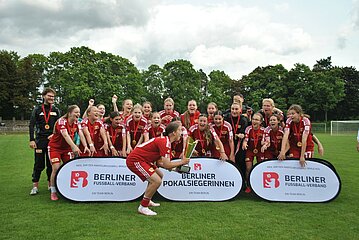 Eine Fußballmannschaft in roten Trikots feiert mit Pokal. Zwei Banner zeigen „Berliner Pokalsiegerinnen“ und „Berliner Fußball-Verband“. Foto: BFV