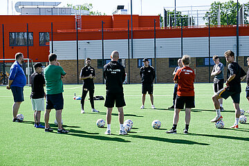 Eine Gruppe steht im Kreis auf einem Fußballplatz und hört einem Trainer zu. Im Hintergrund ist ein Gebäude zu sehen. Foto: Sandra Ritschel
