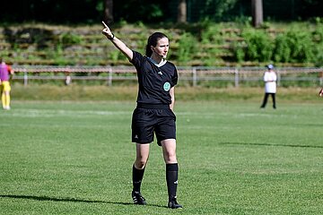 Auf einer grünen Fußballwiese steht eine Schiedsrichterperson in schwarzer Sportkleidung. Sie hebt einen Arm deutlich nach oben und zeigt in eine bestimmte Richtung, als wolle sie eine Entscheidung oder Spielfortsetzung anzeigen. Die andere Hand ruht an der Hüfte. Im Hintergrund sind weitere Spieler*innen und eine Laufbahn sowie eine begrünte Tribüne zu erkennen. Das Wetter wirkt sonnig, und die Szene spielt offensichtlich während eines laufenden Fußballspiels. 