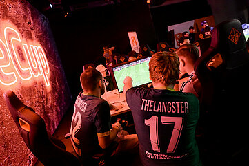 The picture shows a gaming environment with several people sitting at computers and playing a soccer video game. In the foreground, two people can be seen from behind, both wearing dark jerseys with white prints. On the left jersey is the number "12" and a name, on the right jersey the number "17" and the name. Both are sitting in large gaming chairs in front of a monitor on which a soccer pitch can be seen. The scene is bathed in warm, reddish-orange light emanating from large, glowing lettering on the left wall. It reads "eCup" in large letters. In the background, there are other people at similar gaming stations. The atmosphere is reminiscent of an e-sports tournament or gaming event.