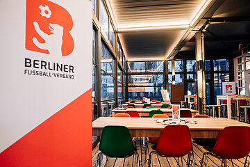 Innenraum mit Tischen und bunten Stühlen; links Logo „Berliner Fußball-Verband“, große Fenster und warme Deckenbeleuchtung.