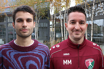 The picture shows two people standing next to each other outdoors. In the background are several birch trees with autumn-colored leaves and a modern building with large glass windows and a grey façade. The person on the left is wearing a knitted sweater in Bordeaux red with an eye-catching pattern of curved lines in blue and purple. A white T-shirt can be seen underneath and a pearl necklace is around his neck. The person on the right is wearing a burgundy sports jacket with a zipper. Several white prints are visible on the jacket: the brand logo "JAKO", the initials "MM" and a club crest with the inscription "Moabiter FSV Berlin" and a stylized figure in motion. Both people are facing the camera.