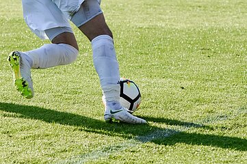 Auf dem Foto ist ein Fußballspieler auf einem grünen Rasenplatz zu sehen. Der Fokus liegt auf den unteren Körperhälften: Der Spieler trägt weiße Shorts, weiße Socken und weiße Fußballschuhe. Er ist gerade dabei, einen schwarz-weißen Fußball zu treten. Die Sonne scheint, was durch die klar erkennbaren Schatten auf dem Rasen sichtbar wird – sowohl vom Ball als auch von den Beinen des Spielers. Der Hintergrund ist nicht sichtbar, sodass der Blick ganz auf die Aktion mit dem Ball gelenkt wird.