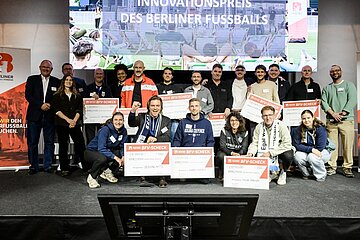 Eine größere Gruppe von Menschen steht auf einer Bühne vor einer großen Leinwand, auf der der Text „Innovationspreis des Berliner Fußballs“ zu lesen ist. Die Personen posieren für ein Gruppenfoto, viele von ihnen halten große symbolische Schecks mit dem Logo des Berliner Fußball-Verbandes in den Händen. Einige tragen Fanartikel wie Schals oder Vereinskleidung. Links und rechts am Rand stehen Roll-Ups mit dem Logo des Verbandes. Der Raum wirkt wie ein Veranstaltungs- oder Kongresssaal, die Stimmung ist festlich und erinnert an eine Preisverleihung oder Ehrung.