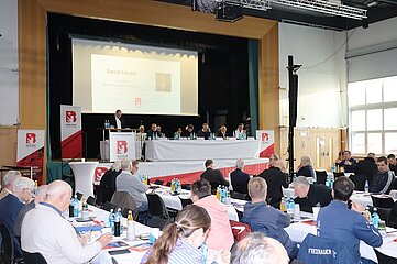 The picture shows a large gathering in a hall that looks like an official conference or election event. In the foreground, numerous people are sitting at long, white-clothed tables arranged in rows. There are documents, brochures and writing materials on the tables. There are also many bottles of water and drinks in shades of blue, green and orange, as well as glasses. In the background is a raised stage with a long table at which several people are seated next to each other. Name tags and several bottles of water can be seen in front of the table. To the left of the stage is a lectern with the logo of the Berlin Football Association (BFV): a red stylized "B" with a soccer motif. The lectern also bears the inscription "BFV-Verbandstag 2025". Behind the stage hangs a large screen onto which a presentation is projected. 