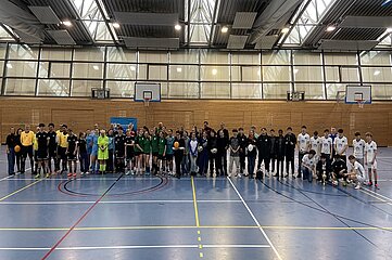Das Foto zeigt eine große Gruppe von Personen, die in einer Sporthalle aufgereiht steht. Die Sporthalle hat einen hohen Deckenbereich mit vielen Fenstern, durch die Tageslicht hineinfällt. Mehrere Basketballkörbe hängen an den Wänden. Die Personen stehen in einer langen Reihe quer durch die Mitte des Bildes. Sie tragen überwiegend Sportkleidung in unterschiedlichen Farben, unter anderem Schwarz, Grün, Blau und Weiß. Einige tragen Trikots, andere Trainingskleidung. Mehrere Personen halten Fußbälle oder Handbälle in den Händen. Die Gruppe wirkt vielfältig: Es sind Menschen unterschiedlicher Körpergrößen und Altersstufen zu sehen. Im Hintergrund ist eine Holzwand zu erkennen, wie sie typischerweise in Sporthallen vorkommt. Auf der linken Seite steht ein blaues Banner, dessen Text im Foto nicht lesbar ist. Der Boden ist ein glatter Hallenboden in Blau mit farbigen Spielfeldmarkierungen in Gelb, Rot und Schwarz.