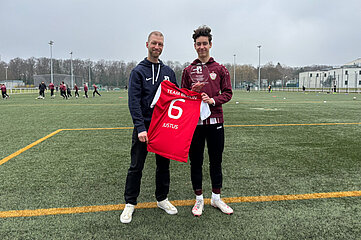 Zwei Personen stehen auf einem Fußballfeld und halten gemeinsam ein rotes Trikot mit der Rückennummer 6 und dem Namen „Justus“ hoch. Eine der Personen hat außerdem eine gläserne Auszeichnung in der Hand. Im Hintergrund sind weitere Spieler beim Training sowie Sportanlagen und umliegende Gebäude zu sehen. 