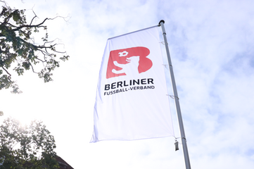 Eine weiße Fahne mit dem Logo des Berliner Fußball-Verbandes weht vor einem blauen Himmel.