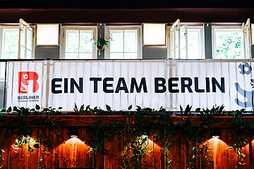 Banner mit Text „Ein Team Berlin“ hängt vor hellen Fenstern, darunter Holzwand mit vielen Pflanzen. Foto: Sandra Ritschel