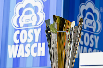 Das Foto zeigt eine Nahaufnahme eines Pokals. Der Pokal ist glänzend und besteht aus goldenen und silbernen Elementen. Er steht im Vordergrund des Bildes und nimmt einen großen Teil des Bildausschnitts ein. Im Hintergrund ist ein Schild zu sehen, das in den Farben Blau und Weiß gestaltet ist. Auf dem Schild steht der Schriftzug „COSY WASCH“. Daneben befindet sich ein stilisiertes Auto, das von einer Wolke umgeben ist – vermutlich das Logo der Firma.
