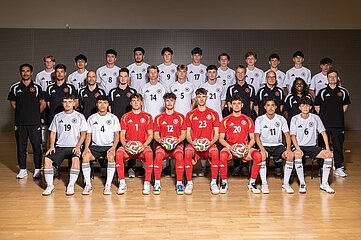 Gruppenfoto einer Futsal-Mannschaft in einer Sporthalle. Insgesamt sind rund 30 Personen in vier Reihen für ein offizielles Teamfoto aufgestellt. Die meisten tragen weiße Trikots mit schwarzen Hosen und weißen Stutzen. Vier Personen in der vorderen Reihe tragen rote Torwarttrikots und halten jeweils einen Futsal-Ball auf dem Schoß. Links und rechts am Rand der Gruppe stehen mehrere Personen in schwarzen Trainingsanzügen oder Poloshirts.  Die Personen sitzen oder stehen dicht beieinander und blicken zur Kamera. Auf den Trikots sind verschiedene Rückennummern zu sehen. Der Hallenboden besteht aus hellem Holz, die Rückwand ist in dunklen Grau- und Brauntönen gehalten. Die Beleuchtung ist gleichmäßig und sorgt für einen professionellen Eindruck. Das Bild vermittelt den Charakter eines offiziellen Mannschafts- oder Kaderfotos vor einem sportlichen Wettbewerb oder Trainingslager.