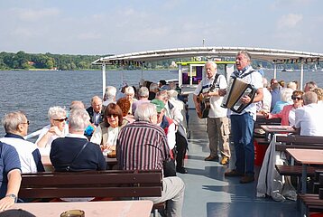 Menschen genießen gemeinsam eine Bootsfahrt, sitzen an Tischen und hören zwei Musikern mit Gitarre und Akkordeon zu.