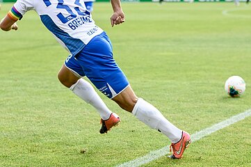 Auf dem Foto ist der untere Körper einer sporttreibenden Person auf einem grünen Fußballrasen zu sehen. Die Person befindet sich in einer Vorwärtsbewegung, offenbar im Sprint. Bekleidet ist sie mit einer blauen kurzen Sporthose, weißen kniehohen Stutzen und orangefarbenen Fußballschuhen. Das weiße Trikot ist nur teilweise sichtbar; auf dem Rücken sind blaue Schriftzüge und eine Rückennummer zu erkennen. Am oberen Arm befindet sich eine Kapitänsbinde mit bunten Regenbogenstreifen. Rechts im Bild liegt ein weißer Fußball mit farbigen Mustern auf dem Rasen, wenige Meter entfernt von der laufenden Person. Weiße Spielfeldlinien markieren den Boden. Der Hintergrund ist unscharf und zeigt ausschließlich weitere Rasenfläche, ohne erkennbare Zuschauende oder Gebäude. Das Bild vermittelt eine sportliche Spielsituation im Freien während eines Fußballspiels oder Trainings.
