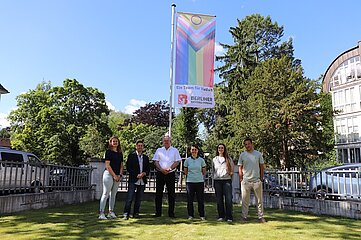 Das Foto zeigt sechs Personen, die gemeinsam auf einer grünen Rasenfläche stehen. Sie sind in lockerer Formation nebeneinander aufgestellt und blicken in Richtung Kamera. Hinter ihnen ist eine große, bunte Fahne zu sehen. Auf der Fahne steht in klar lesbarer Schrift: „Ein Team für Vielfalt“, begleitet vom Logo des Berliner Fußball-Verbandes. Die Farben der Fahne sind lebendig und symbolisieren Vielfalt. Im Hintergrund erkennt man Bäume und Gebäude, die auf eine städtische Umgebung hinweisen. Die Szene wirkt freundlich und offen. Es sind keine sichtbaren Barrieren vorhanden, und die Darstellung legt Wert auf Gemeinschaft und Inklusion.