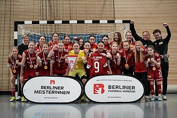 Eine Mädchen- oder Frauenfußballmannschaft posiert jubelnd in einer Sporthalle vor einem Hallenfußballtor. Alle tragen einheitliche rote Trikots des 1. FC Union Berlin. Einige Teammitglieder halten stolz Medaillen in die Kamera, andere zeigen Siegergesten wie erhobene Arme oder Daumen nach oben. In der Mitte steht eine Person im gelben Torwarttrikot, die ein Wimpelbanner mit dem Logo des Berliner Fußball-Verbands präsentiert. Links und rechts im Vordergrund befinden sich zwei ovale Werbetafeln. Die linke zeigt den Schriftzug „Berliner Meisterinnen – Ein Team Berlin“, die rechte „Berliner Fußball-Verband – Ein Team Berlin“, jeweils mit rotem Logo. Die Gruppe wirkt ausgelassen und feierlich, als würde sie gemeinsam einen Meistertitel oder Turniersieg feiern. Die Umgebung deutet auf eine typische Indoor-Sporthalle hin, mit Holzwandverkleidung und einem engmaschigen Netz hinter dem Tor.