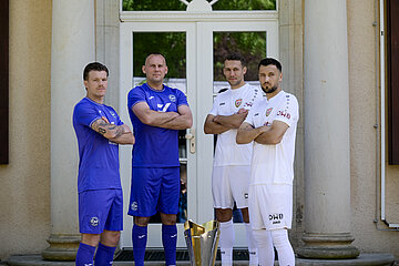 Vor einem Gebäude mit Säulen und einer Glastür stehen vier Personen. Zwei von ihnen tragen blaue Sportkleidung, die anderen beiden weiße. Alle vier Personen stehen mit verschränkten Armen um einen großen Pokal, der mittig auf dem Boden platziert ist. Die Gesichter der Personen sind unkenntlich gemacht. Im Hintergrund ist die Architektur des Gebäudes sichtbar, es sind jedoch keine weiteren Details oder Texte zu erkennen. Foto: Sandra Ritschel