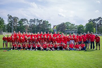 Das Foto zeigt eine große Gruppe von Menschen unterschiedlichen Alters, Geschlechts und Erscheinungsbildes, die gemeinsam auf einer grünen Rasenfläche stehen oder sitzen. Alle tragen rote T-Shirts, was auf eine gemeinsame Zugehörigkeit zu einem Team oder einer Veranstaltung hinweist – möglicherweise ein Sportevent. Einige Personen sitzen auf dem Boden, andere stehen. Die Gruppe wirkt fröhlich und verbunden. Im Hintergrund sind grüne Bäume und einige Gebäude zu sehen. Der Himmel ist leicht bewölkt, aber es scheint ein sonniger Tag zu sein.