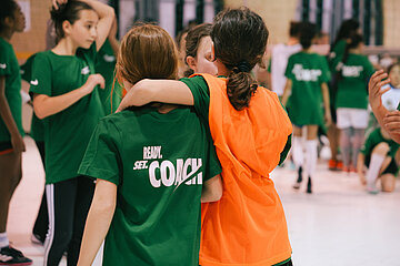 Kinder trainieren in einer Halle; zwei Kinder stehen im Vordergrund eng nebeneinander, umgeben von weiteren spielenden Kindern in grünen Shirts. Foto: Lea Gleisberg