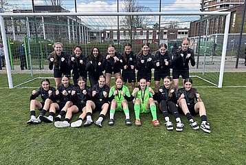 Vor einem Fußballtor posiert eine komplette Mädchenmannschaft geschlossen und gut gelaunt. Die Spielerinnen tragen schwarze Trainingsjacken, zwei in der Mitte sitzen in leuchtend grünen Torwarttrikots. Alle haben die Fäuste motiviert nach vorne gestreckt, als würden sie gemeinsam jubeln oder Teamgeist zeigen. Die Gruppe sitzt und steht auf einem gepflegten Rasenplatz, im Hintergrund sind ein Zaun, Trainingsfelder und einige Gebäude zu sehen. Die Stimmung wirkt sportlich, stolz und voller Zusammenhalt.