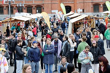 Das Foto zeigt eine belebte Szene auf einem offenen Markt oder Straßenfest. Zahlreiche Menschen bewegen sich zwischen mehreren Verkaufsständen, die aus Holzrahmen bestehen und mit Stoffdächern überspannt sind. Einige Personen unterhalten sich, andere betrachten die ausgestellten Waren oder benutzen ihre Mobiltelefone. Die Menschen sind unterschiedlich gekleidet, was auf eine vielfältige Besuchergruppe hinweist.  Im Hintergrund steht ein großes Gebäude aus roten Ziegelsteinen mit mehreren hohen, rundbogigen Fenstern und Türen. Über dem Markt sind verschiedene Banner und Schilder aufgehängt, die auf Angebote oder Veranstaltungen hinweisen könnten. Die Atmosphäre wirkt lebendig und einladend.