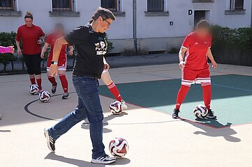 Das Foto zeigt eine Gruppe von mehreren Personen auf einer hellen, ebenen Spielfläche im Freien, die mit Linien markiert ist. Jede sichtbare Person hat einen Fußball in der Nähe der Füße; mehrere Bälle liegen gleichmäßig verteilt auf dem Boden. Die Personen bewegen sich gleichzeitig in dieselbe Richtung, was auf eine gemeinsame Übung hindeutet. Im Vordergrund links steht eine Person in Alltagskleidung mit schwarzem Oberteil, Jeans und Sportschuhen. Der Blick und die Körperhaltung sind auf den Fußball am Boden ausgerichtet, ein Fuß ist leicht angehoben, als würde der Ball kontrolliert oder vorwärts bewegt. Im Hintergrund und rechts stehen weitere Personen in roter Sportkleidung mit weißen Streifen, darunter Trikots, Shorts und kniehohe Stutzen. Auch diese Personen führen jeweils einen Ball mit den Füßen. Im Hintergrund ist ein helles Gebäude mit mehreren Fenstern zu sehen, einige davon mit Gitterstäben versehen. Ein Metallzaun und niedrige grüne Büsche grenzen die Spielfläche vom Gebäude ab. An einer Wand ist der Buchstabe „D“ angebracht. Das Licht ist hell und gleichmäßig, was auf sonniges Wetter schließen lässt. Die Gesichter aller Personen sind unscharf dargestellt, sodass keine Identifizierung möglich ist. Die Szene vermittelt den Eindruck eines gemeinsamen Fußballtrainings oder einer angeleiteten Übung, bei der mehrere Personen gleichzeitig und koordiniert mit dem Ball arbeiten.
