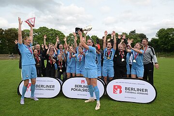 Eine Gruppe von Fußballspielern und Betreuern feiert auf einem grünen Rasenplatz einen Sieg. Die Spieler tragen hellblaue Trikots, einige halten einen Pokal in die Höhe. Vor der Gruppe stehen drei Banner mit der Aufschrift „BERLINER FUSSBALL-VERBAND“ und „BERLINER POKALSIEGER“. Im Hintergrund sind Bäume und ein bewölkter Himmel zu sehen.